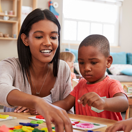 A photographic style image of a child care worker. She's helping a young boy with an educational style activity.